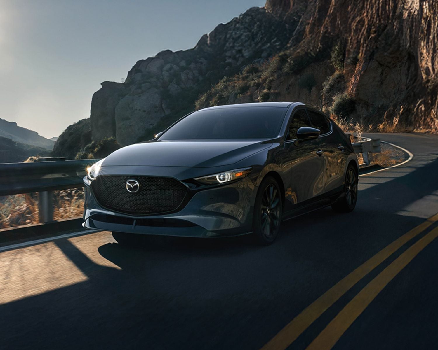Front 3/4 shot of the 2026 MAZDA3 Hatchback driving down winding road.